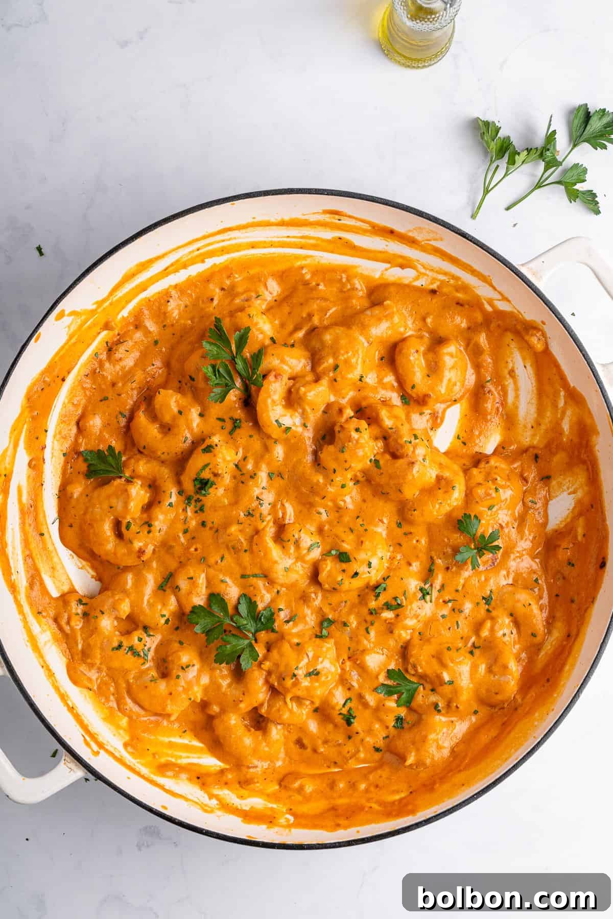 A pristine white pan brimming with creamy shrimp in a rich, inviting sauce, beautifully garnished with vibrant fresh parsley. The delectable dish is set on a light marble surface, with a subtle sprig of parsley and an elegant bottle of olive oil forming a tasteful backdrop.