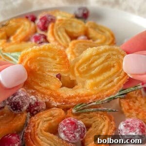 A close-up of a hand holding a heart-shaped Puff Pastry Cookie, with more cookies, sugared cranberries, and rosemary arranged on a plate in the background.