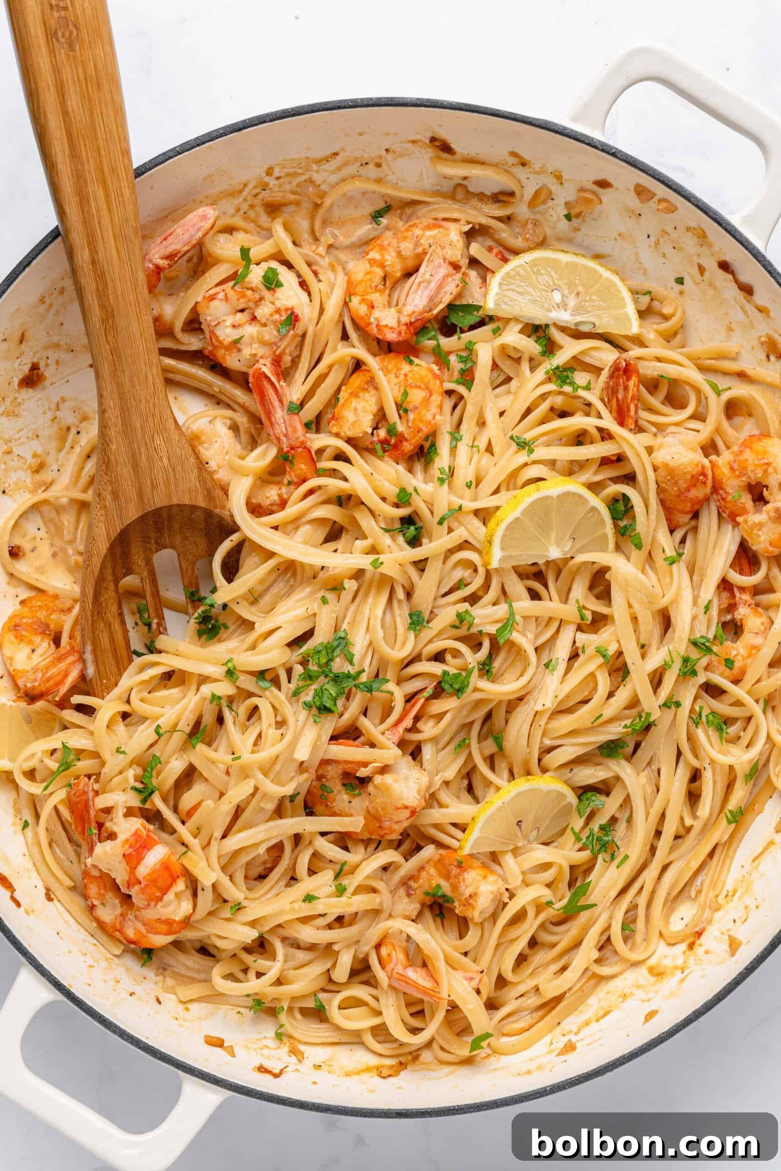 Zesty Lemon Garlic Shrimp Pasta 2 A white pan filled with creamy shrimp linguine garnished with fresh parsley and lemon slices. A wooden spoon is placed within the pasta, and the dish sits on a light surface.