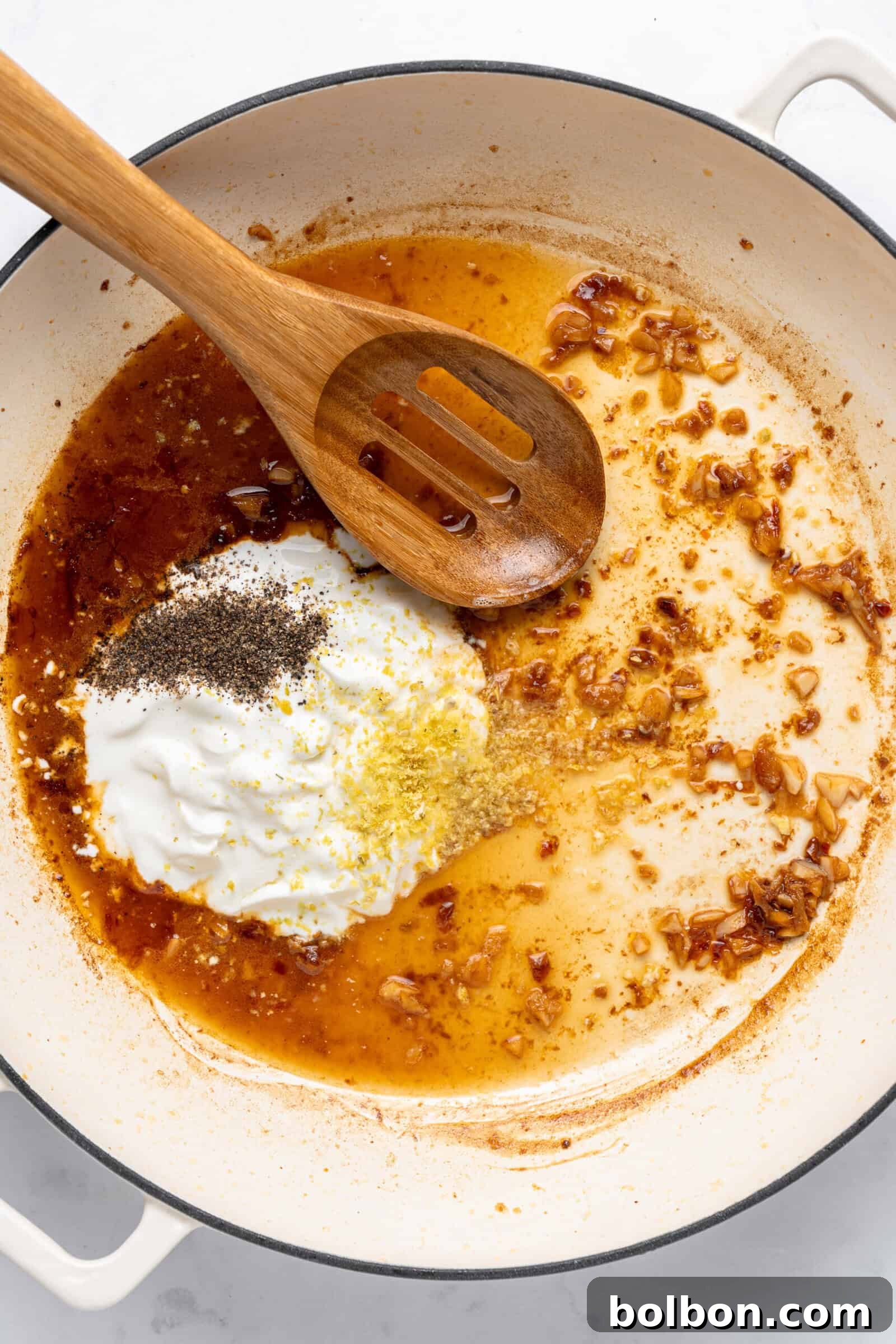 Zesty Lemon Garlic Shrimp Pasta 6 A white pan containing a wooden spoon, sautéed onions, seasonings, and a dollop of yogurt being mixed. The spices include black pepper and lemon zest, creating a rich, aromatic sauce base.
