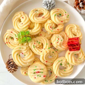 A white plate filled with Shortbread Butter Cookies topped with red and green sprinkles, surrounded by pinecones and small wrapped gifts, creates a festive holiday display.