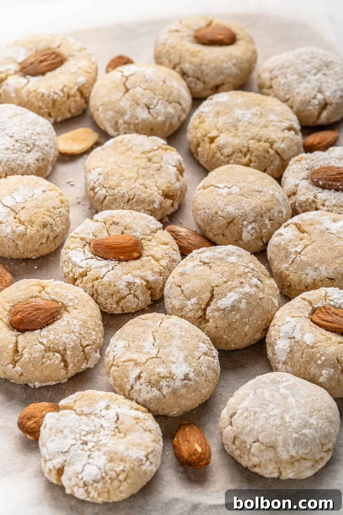 Almond Amaretti: Italian Cookie Tradition 11 A batch of Italian Amaretti Cookies dusted with powdered sugar is arranged on baking paper. Some cookies have a whole almond on top, while others are plain. A few loose almonds are scattered among the cookies.
