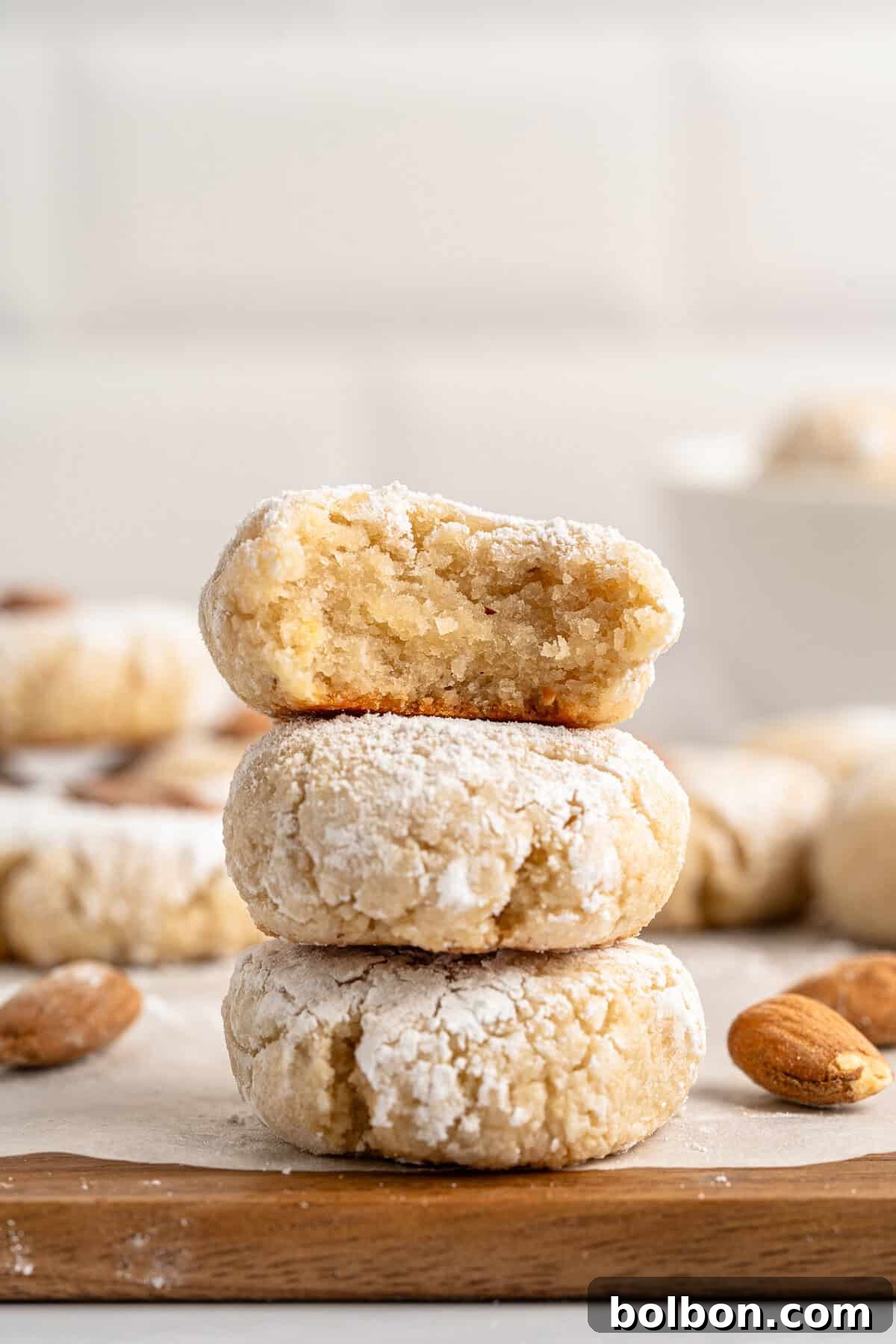 Almond Amaretti: Italian Cookie Tradition 12 A stack of three powdered Italian Amaretti cookies on a wooden surface. The top cookie has a bite taken out, revealing a soft interior. Whole almonds are scattered around, with more cookies blurred in the background.