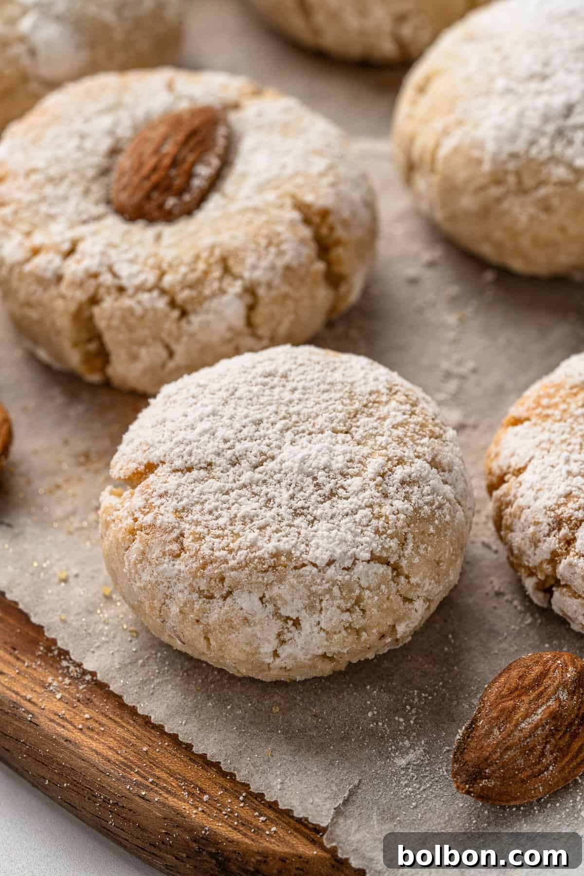 Almond Amaretti: Italian Cookie Tradition 13 Close-up of freshly baked almond Italian Amaretti cookies dusted with powdered sugar, placed on parchment paper with whole almonds on top of each cookie, resting on a wooden surface.