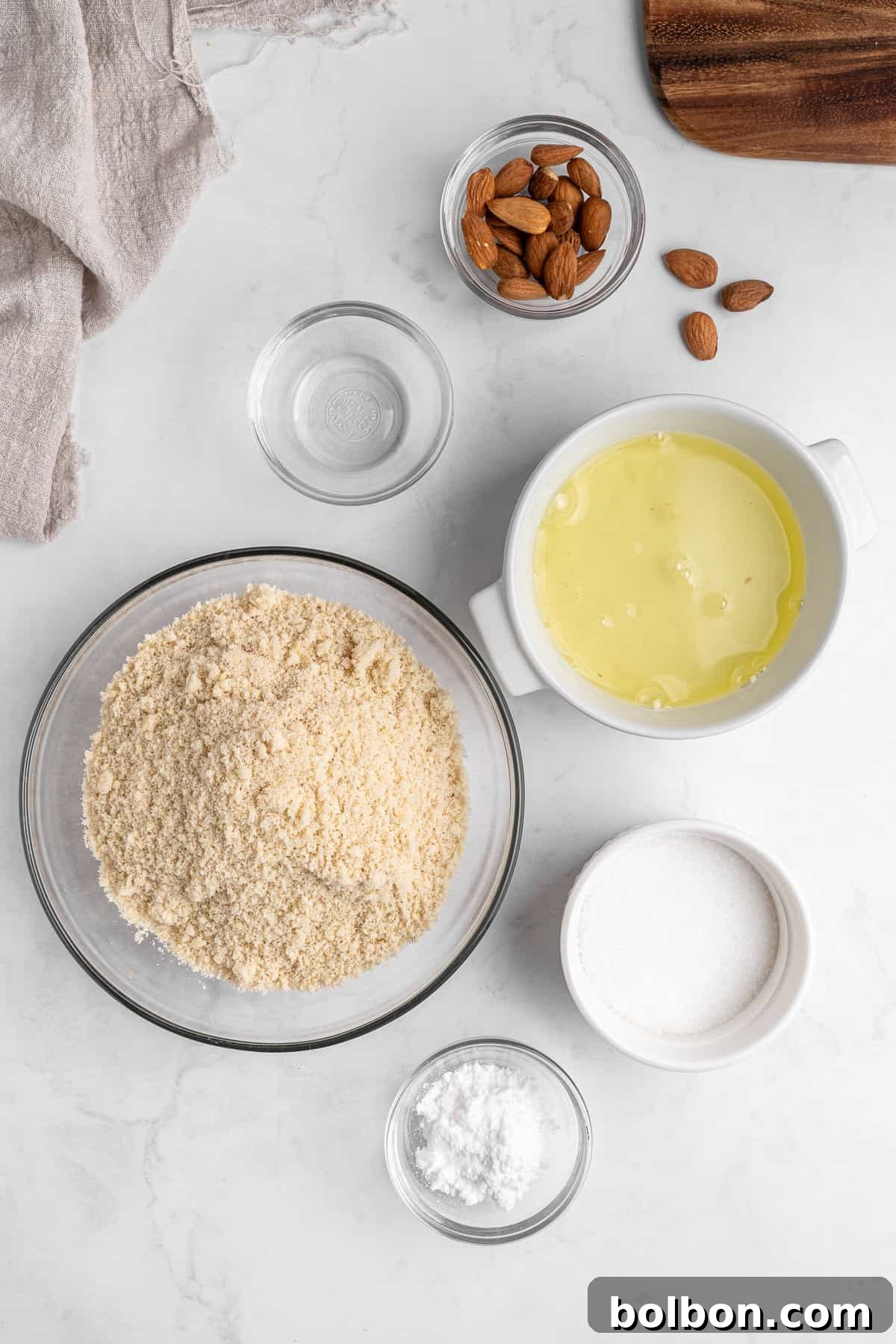 Almond Amaretti: Italian Cookie Tradition 3 Top-down view of baking ingredients on a white surface, including a bowl of almond flour, a bowl with egg whites, a bowl of whole almonds, a small bowl of powdered sugar, and a glass of water, with a beige cloth nearby.