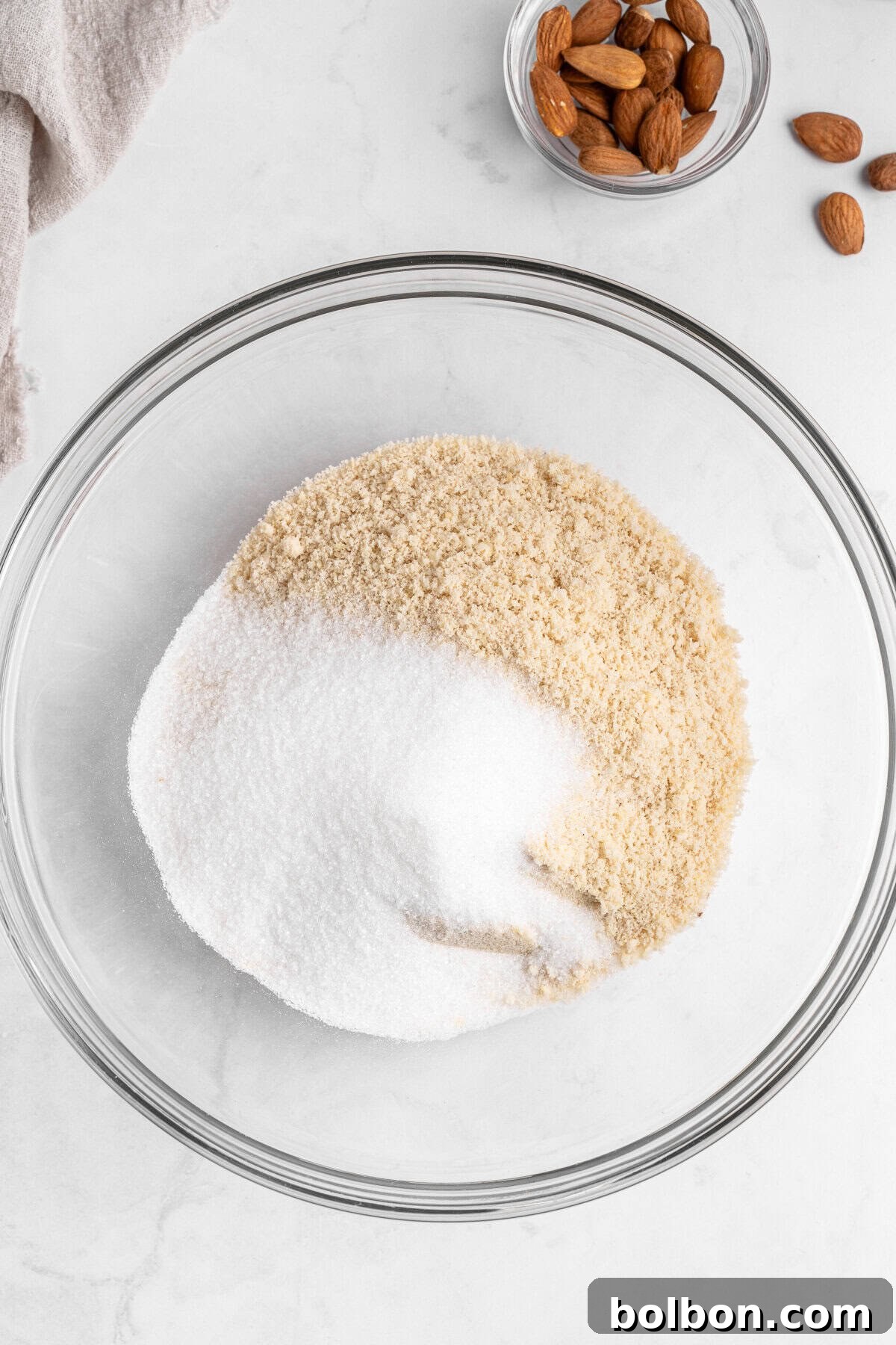 Almond Amaretti: Italian Cookie Tradition 4 A glass bowl containing white and brown sugar is placed on a marble surface. To the top right, there's a small bowl filled with almonds, and a few almonds are scattered nearby. A beige cloth is partially visible to the top left.