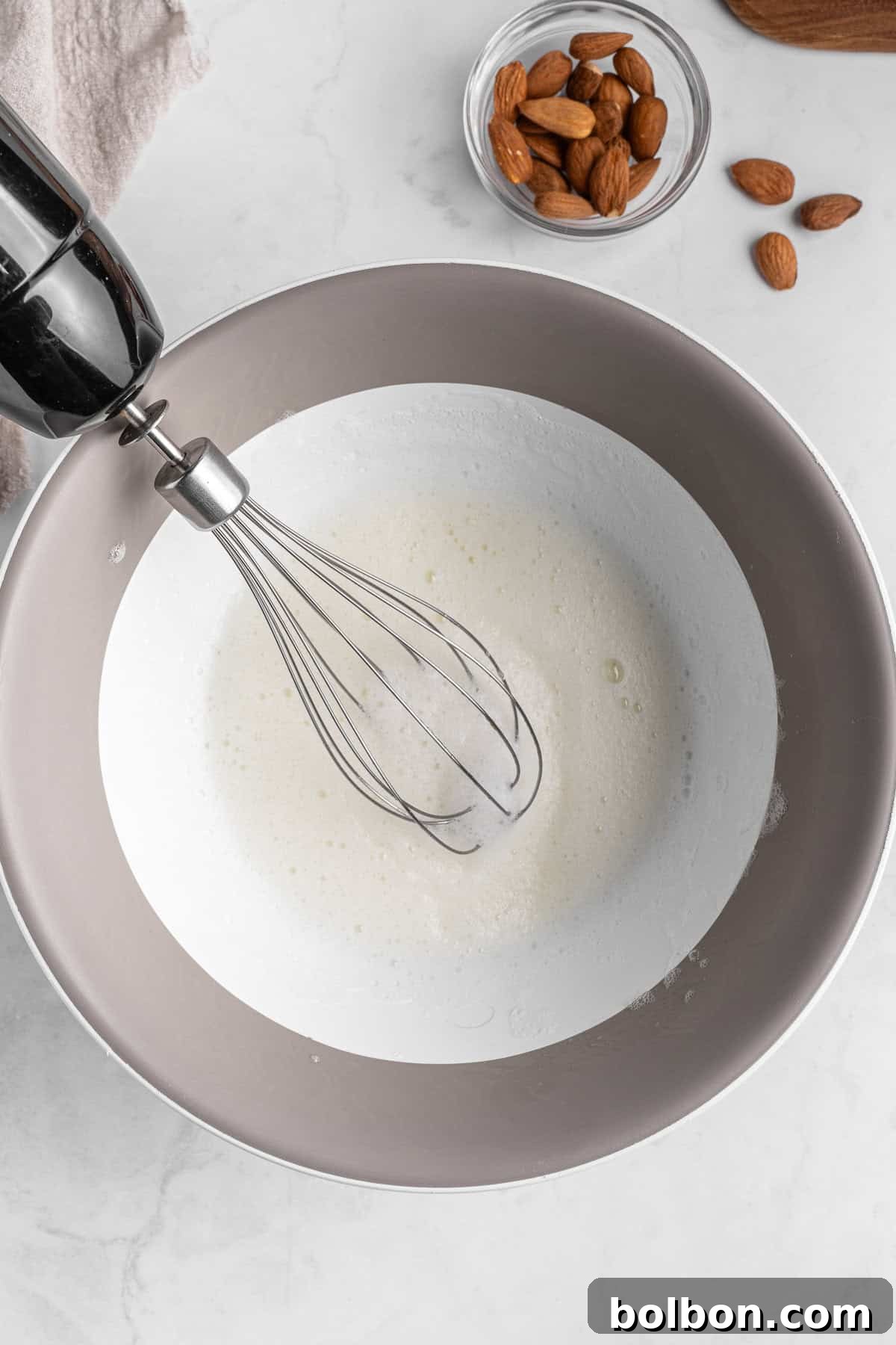 Almond Amaretti: Italian Cookie Tradition 5 A whisk attached to an electric mixer is beating egg whites in a mixing bowl. Beside the bowl, there is a small glass bowl filled with almonds on a marble countertop.