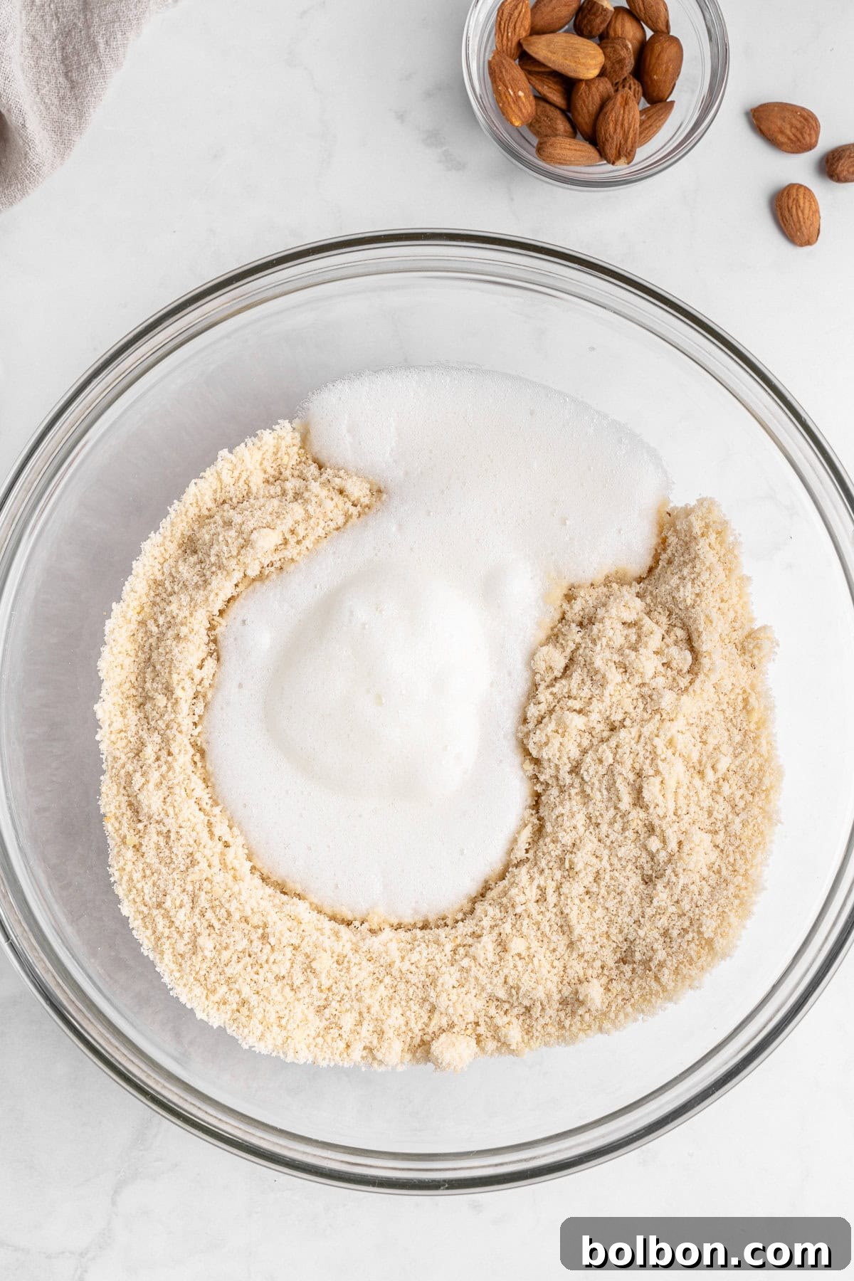 Almond Amaretti: Italian Cookie Tradition 6 A mixing bowl contains almond flour and foamy egg whites, partially blended together. A small bowl with whole almonds and a few loose almonds are visible in the top right corner on a light-colored marble surface.
