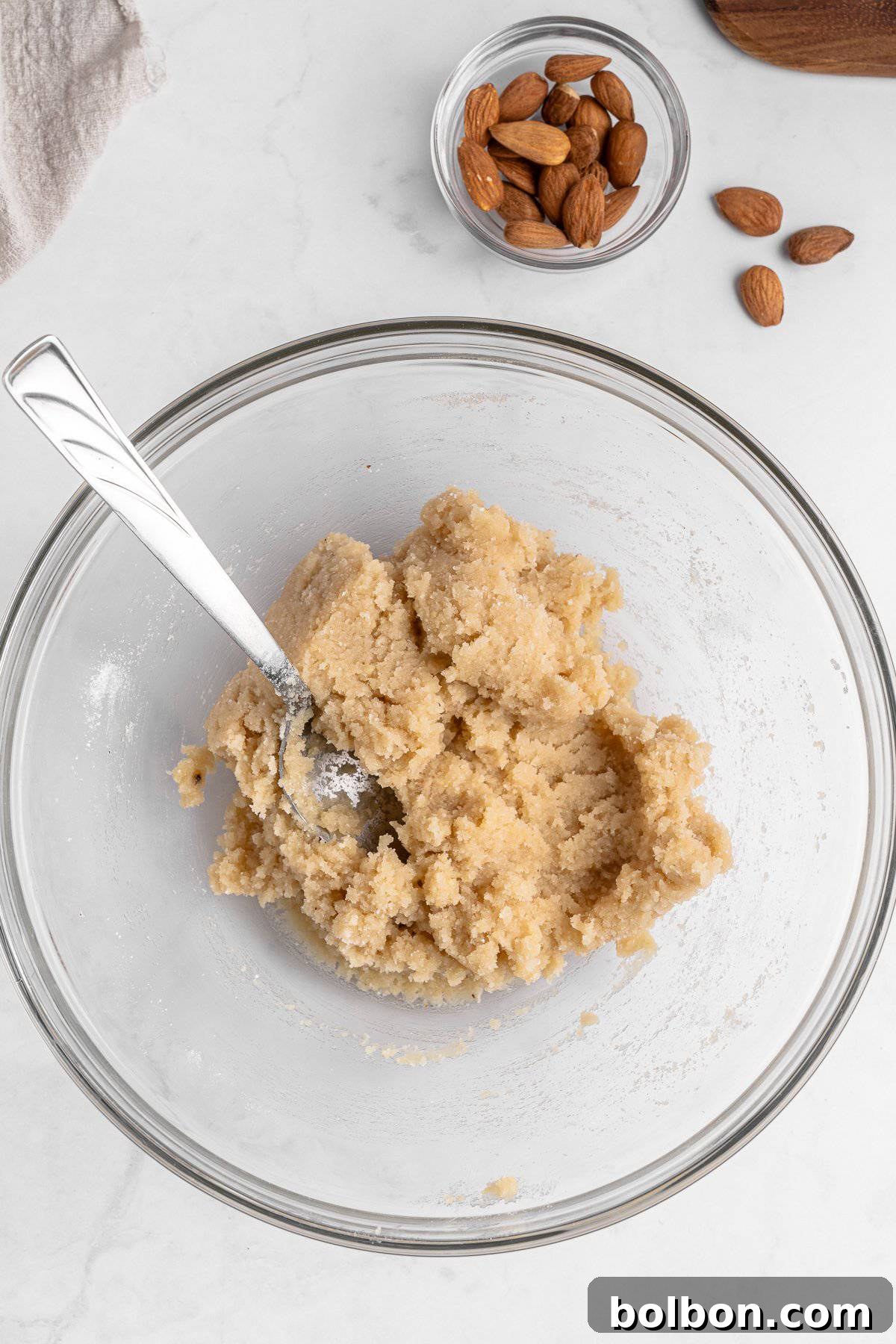 Almond Amaretti: Italian Cookie Tradition 7 A glass bowl on a marble surface containing mixed almond paste with a spoon in it. A small glass bowl with whole almonds and a few scattered almonds are in the background.