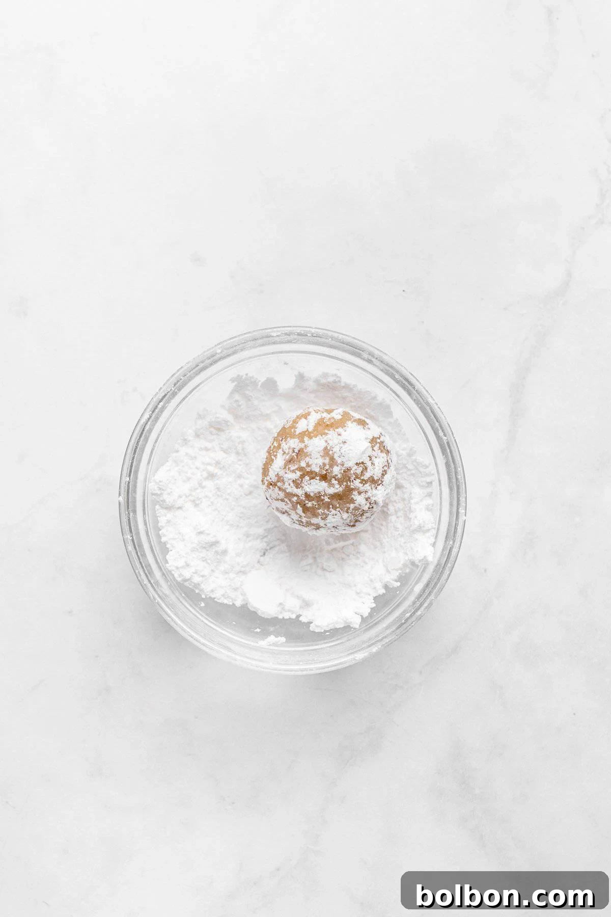 Almond Amaretti: Italian Cookie Tradition 8 A small round ball of cookie dough sits in a clear glass bowl filled with powdered sugar, placed on a light marble surface.
