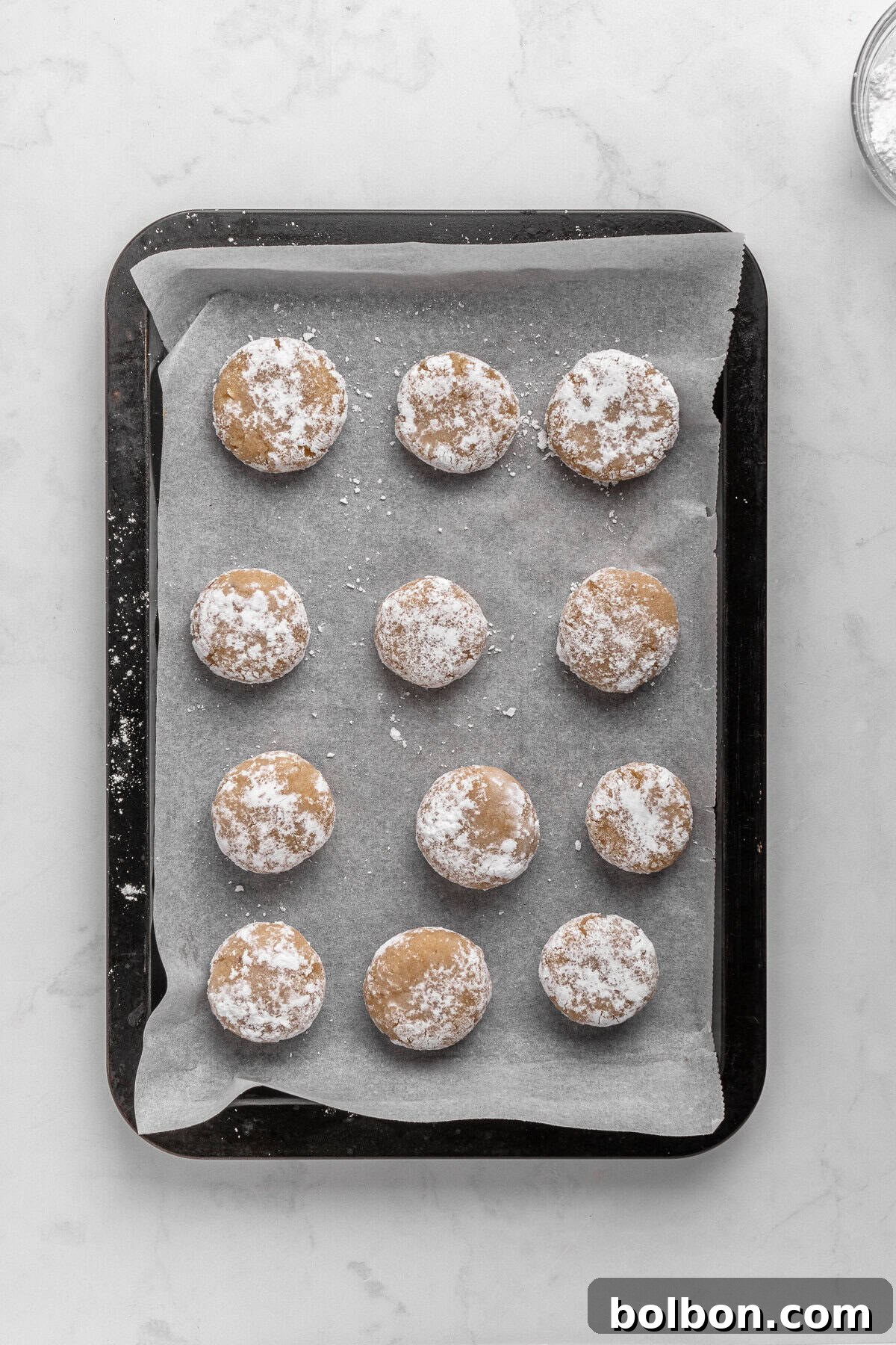 Almond Amaretti: Italian Cookie Tradition 9 A baking tray with twelve raw, round cookies covered in powdered sugar. The cookies are arranged in four rows on parchment paper, ready for baking. The background is a white, textured surface.