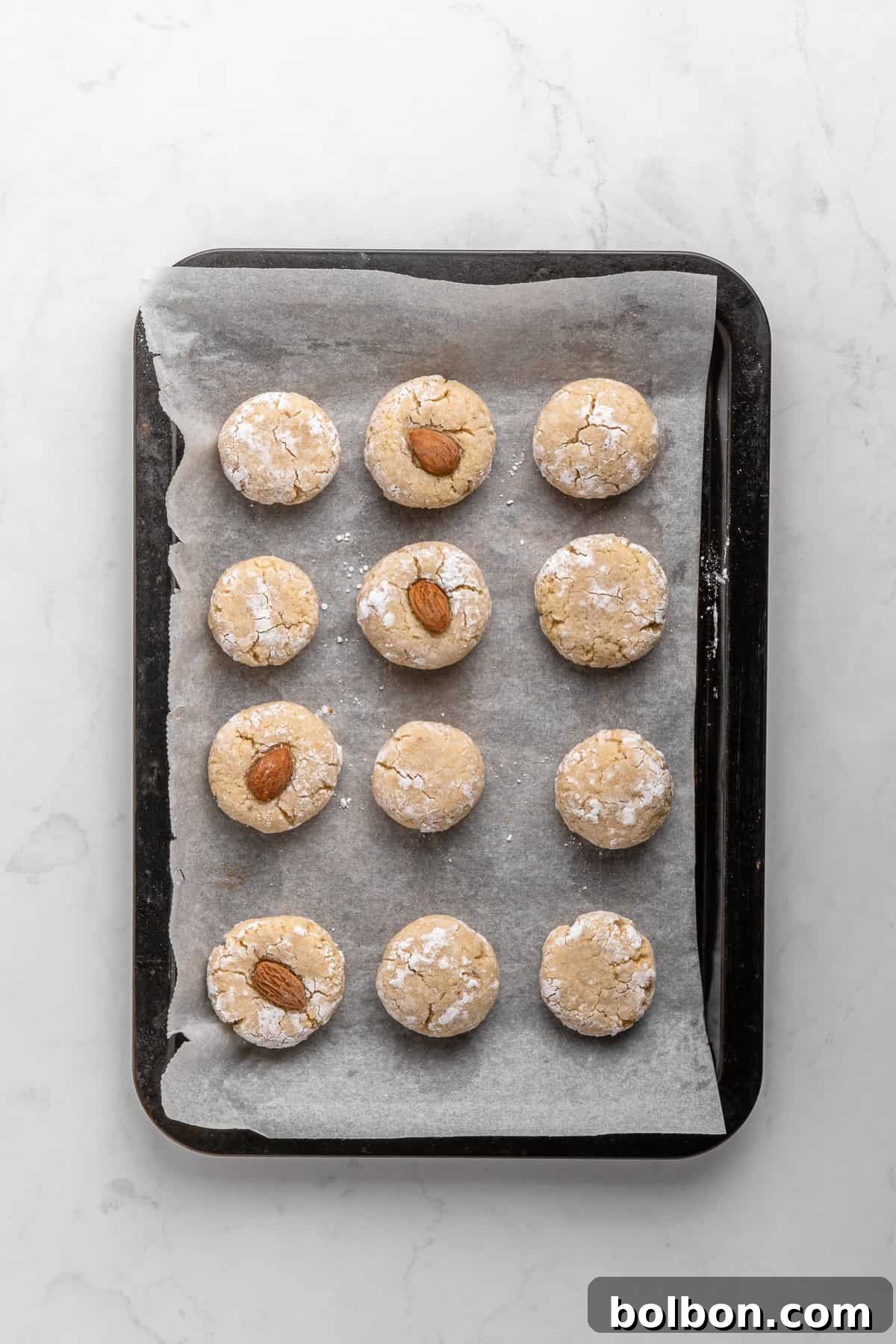 Almond Amaretti: Italian Cookie Tradition 10 A baking tray lined with parchment paper holds twelve Italian Amaretti cookies, arranged in a grid. Some cookies have a whole almond on top. The tray rests on a white marble surface.