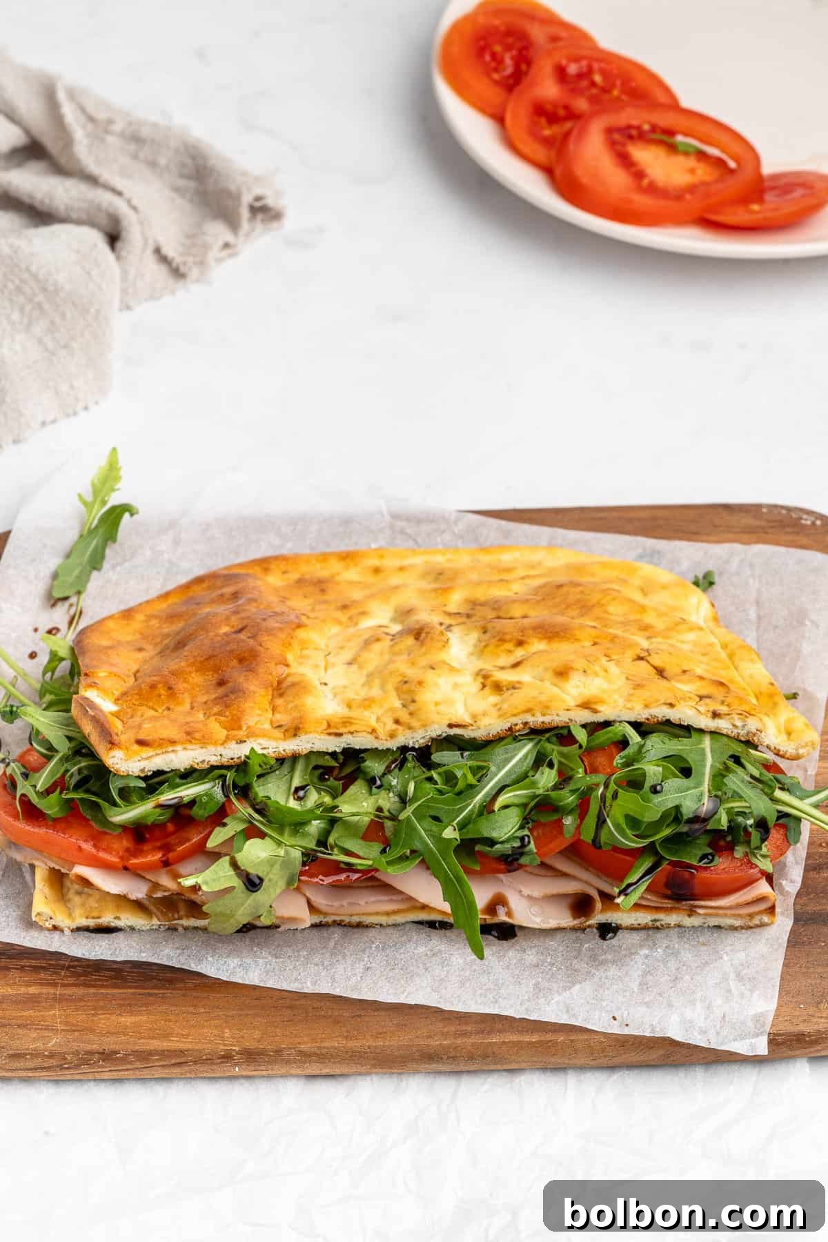 Protein-Packed Cottage Cheese Flatbread 2 A sandwich made with focaccia bread, filled with turkey slices, fresh tomatoes, and arugula, sits on parchment paper on a wooden cutting board. A plate with sliced tomatoes is in the background, next to a beige cloth.