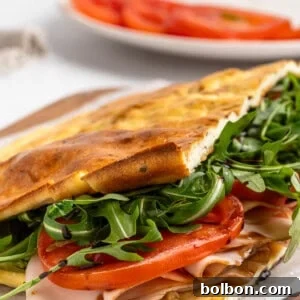 Protein-Packed Cottage Cheese Flatbread 11 A freshly made sandwich on a wooden board, filled with sliced turkey, fresh arugula, and tomato slices. The sandwich is on ciabatta bread, slightly toasted. A white plate with additional tomato slices is blurred in the background.