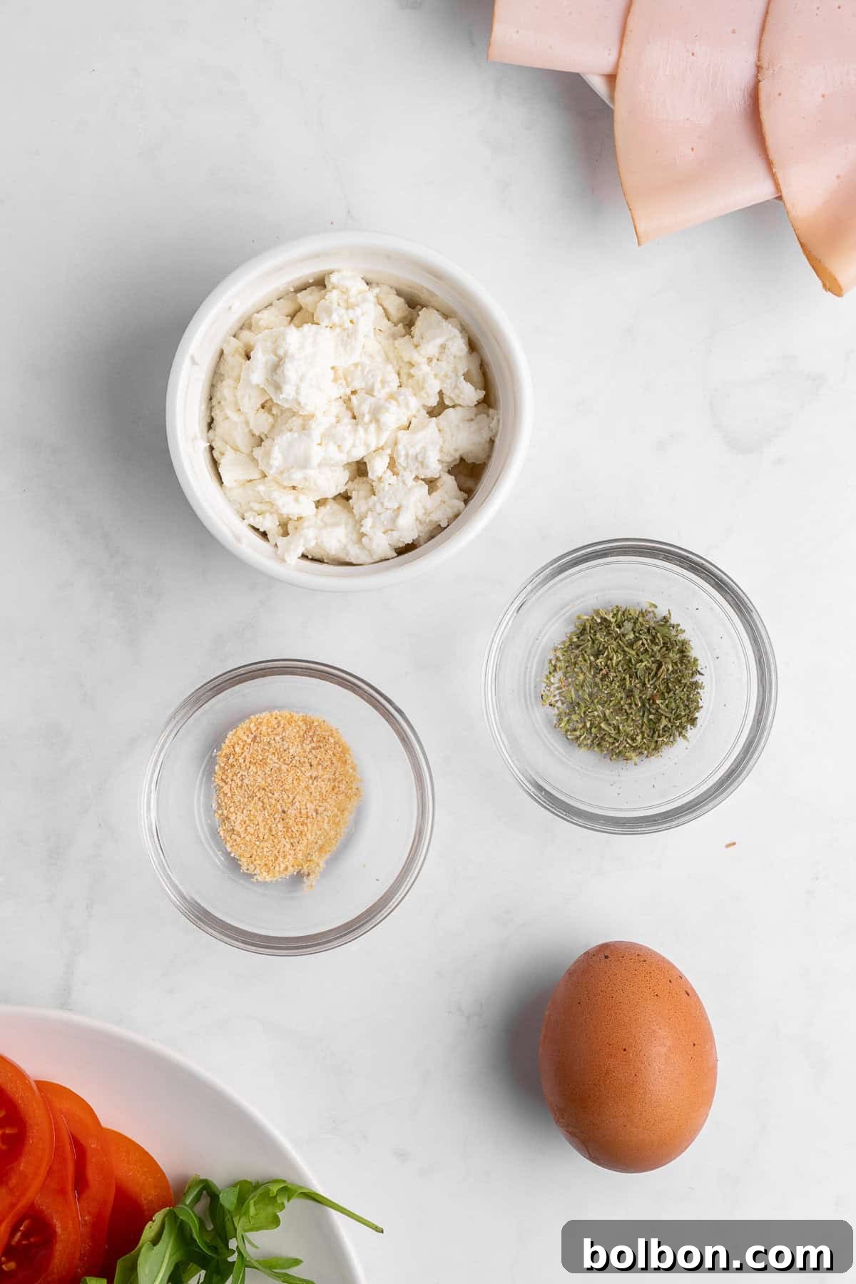 Protein-Packed Cottage Cheese Flatbread 3 Top-down view of ingredients on a white surface: a bowl of ricotta cheese, dried herbs, garlic powder, sliced ham, a brown egg, fresh tomato slices, and arugula leaves.