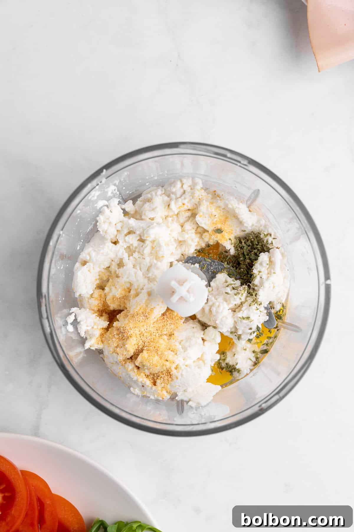 Protein-Packed Cottage Cheese Flatbread 4 A food processor filled with ricotta cheese, breadcrumbs, herbs, and seasoning. Below, a plate with sliced tomatoes and basil is partially visible. The background is a white marble surface.