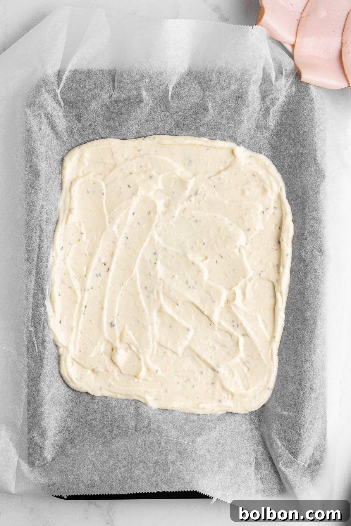 Protein-Packed Cottage Cheese Flatbread 6 A baking tray lined with parchment paper holds an even spread of creamy, seasoned dough or batter. The surface is smooth and speckled with herbs. A few slices of deli meat are visible in the corner on a light background.
