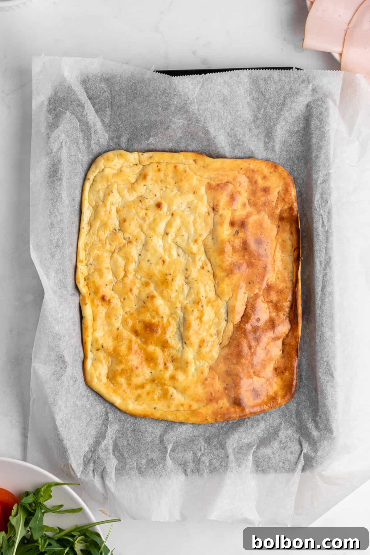 Protein-Packed Cottage Cheese Flatbread 7 A freshly baked rectangular cottage cheese flatbread on parchment paper, resting on a marble surface. Ingredients like arugula, tomatoes, and slices of ham are partially visible around the quiche.