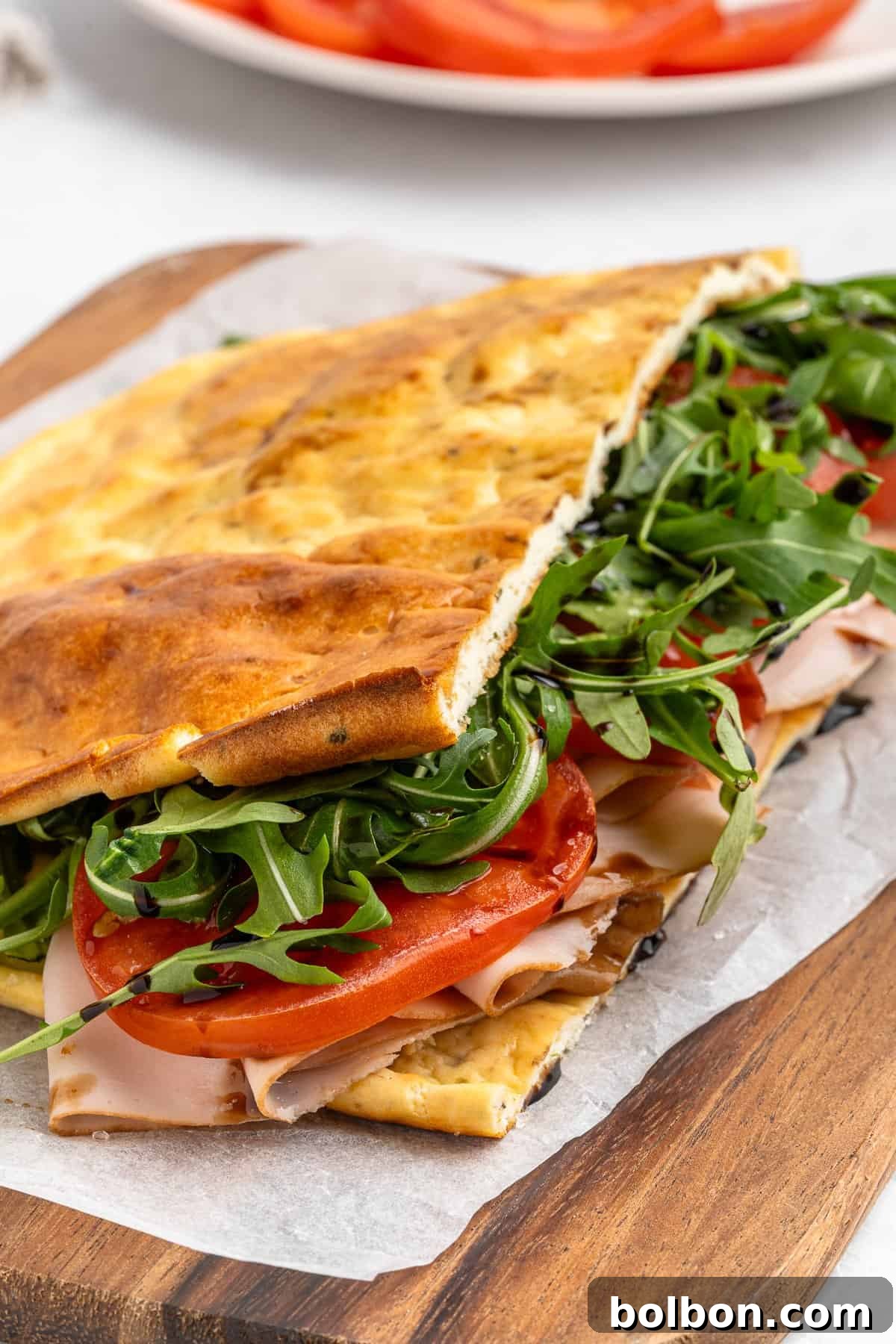 Protein-Packed Cottage Cheese Flatbread 9 A sandwich on a wooden board, made with flatbread, filled with arugula, sliced tomatoes, and deli turkey. The flatbread is folded over the ingredients, which are visible. A plate with more tomato slices is in the background.