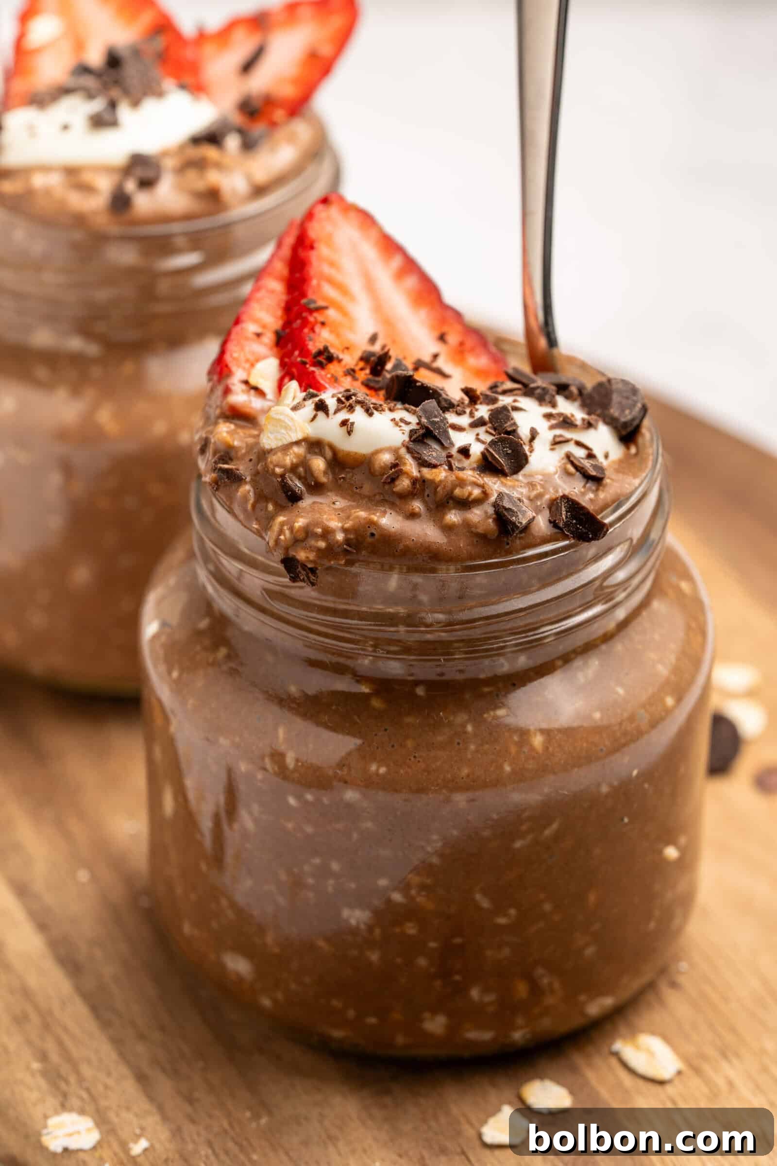 A small glass jar filled with chocolate overnight oats, topped with sliced strawberries, whipped cream, chocolate shavings, and oats. A spoon is inserted into the jar. Another similar jar is in the background.