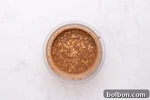 A glass bowl filled with a mixture of oats and chocolate liquid sits on a light marble surface.