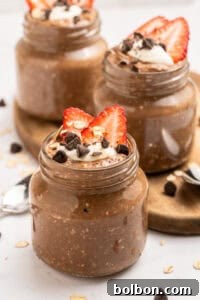 Three jars of chocolate overnight oats are topped with sliced strawberries, chocolate chips, oats, and a dollop of yogurt or cream. The jars sit on a wooden tray with spoons nearby.