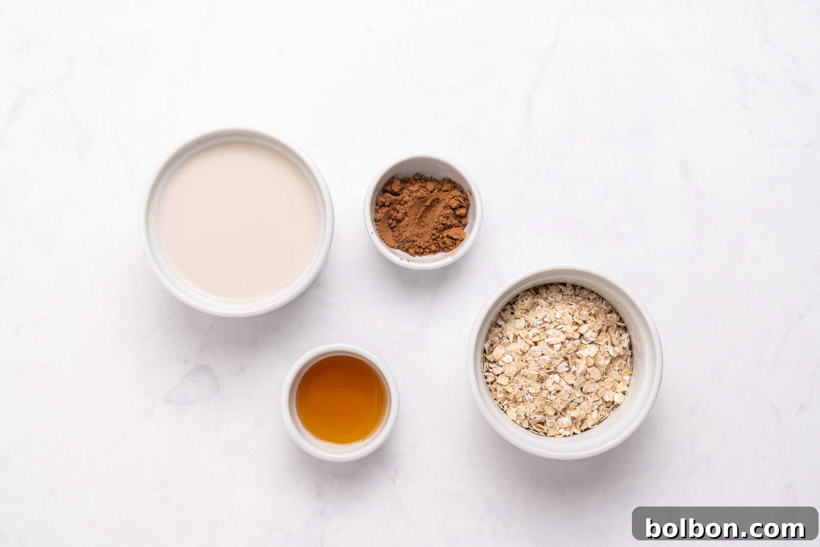 Four small bowls on a white surface containing oat milk, cocoa powder, rolled oats, and maple syrup, arranged in a flat lay style.