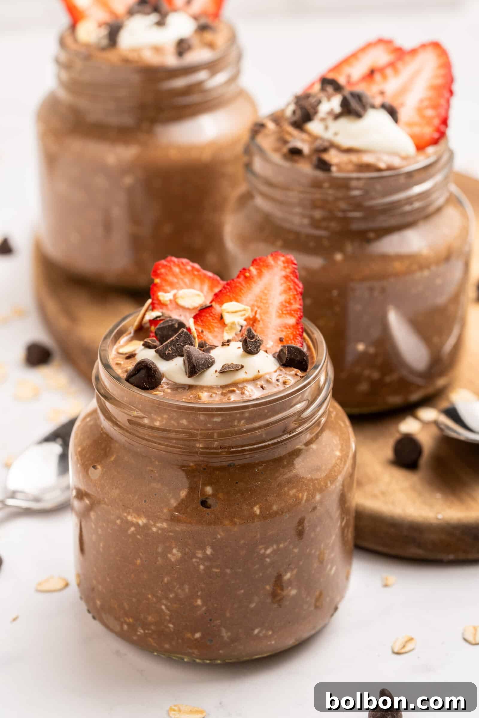 Three jars of chocolate overnight oats are topped with sliced strawberries, chocolate chips, oats, and a dollop of yogurt or cream. The jars sit on a wooden tray with spoons nearby.