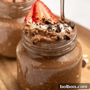 A small glass jar filled with chocolate overnight oats, topped with sliced strawberries, whipped cream, chocolate shavings, and oats. A spoon is inserted into the jar. Another similar jar is in the background.