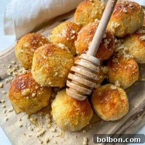 Golden Honey Almond Delights 3 A pile of golden baked pastry balls on a wooden board, drizzled with honey using a honey dipper and sprinkled with crushed nuts, evokes the delightful look of Honey Almond Cookies, with a white cloth in the background.
