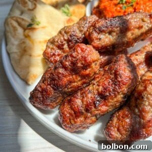 Grilled minced meat kebabs, known as cevapi, served with flatbread, red sauce, and sliced onions on a white plate, all placed on a light wooden surface.