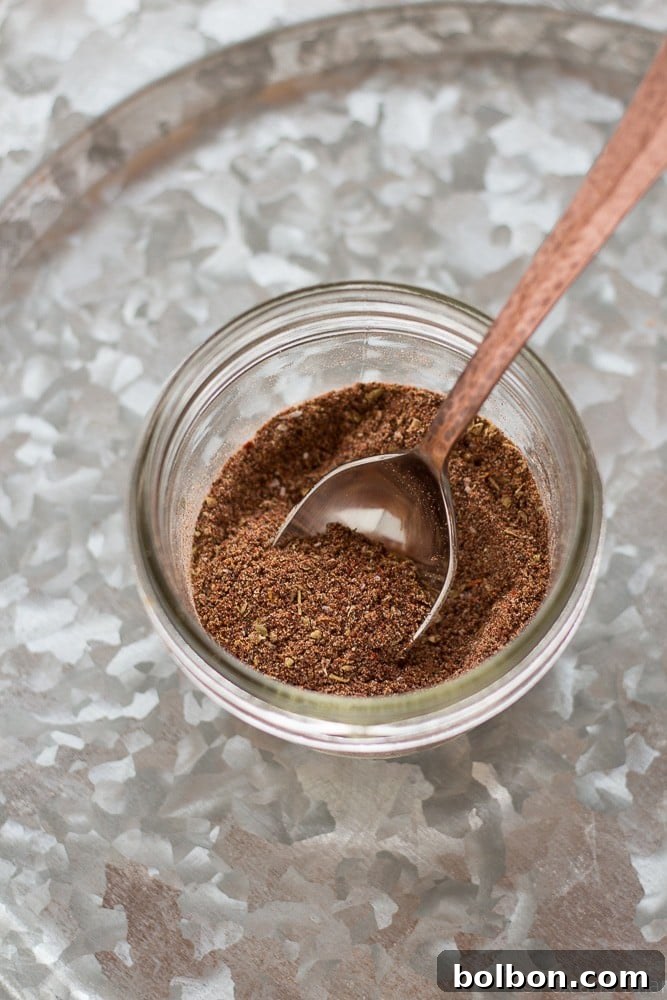 A mason jar filled with homemade taco seasoning stands on a wooden surface, with cooking ground beef in the background, illustrating readiness for use in dinner preparation.