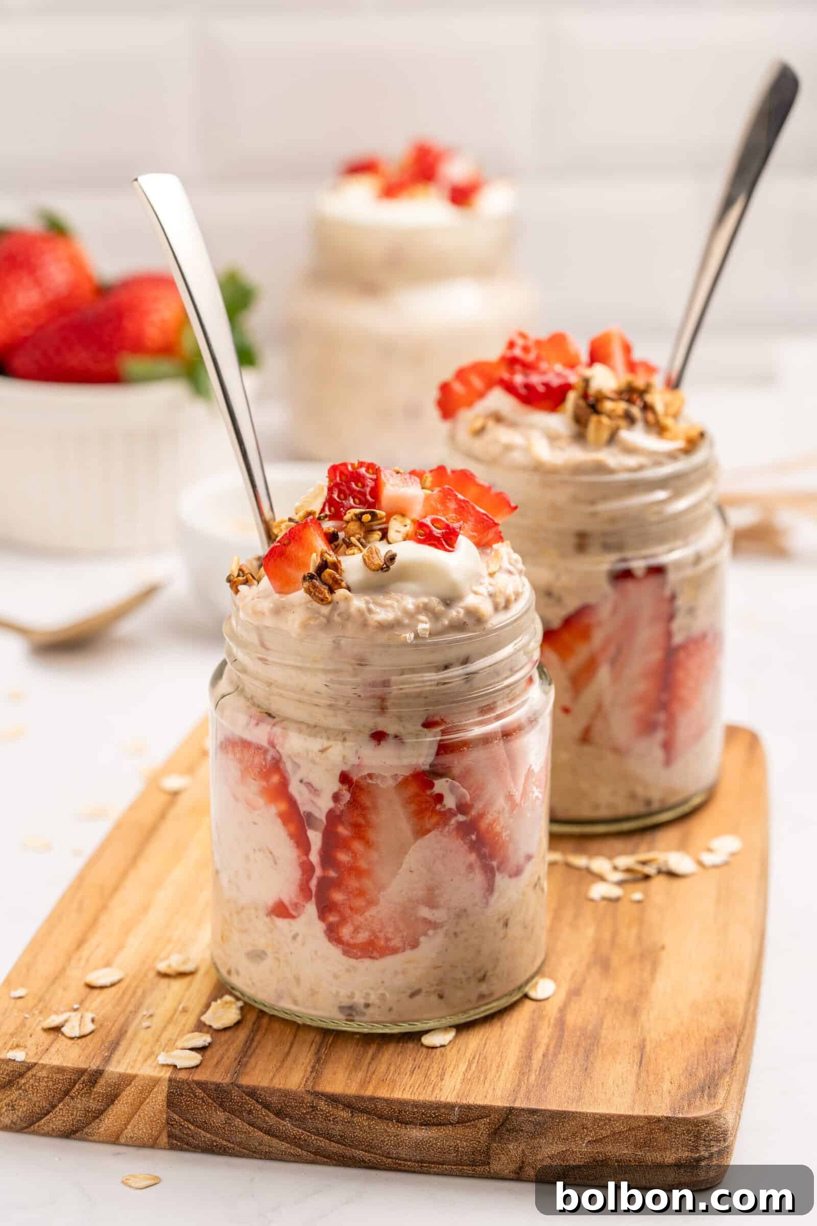 Two glass jars filled and layered with sliced strawberries and topped with granola and yogurt, sit on a wooden board. A bowl of strawberries is in the background, and oats are scattered around.