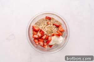 A glass bowl filled with rolled oats, chopped fresh strawberries, and a dollop of yogurt, placed on a light marble surface.