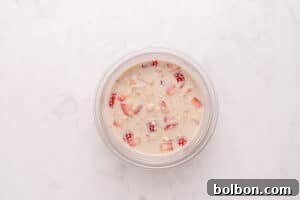 A glass bowl filled with milk, oats, and chopped strawberries sits on a light, marble surface.