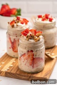 Three glass jars filled with overnight oats, layered with fresh strawberry slices and topped with granola and strawberry pieces, are arranged on a wooden board. A bowl of whole strawberries is in the background.