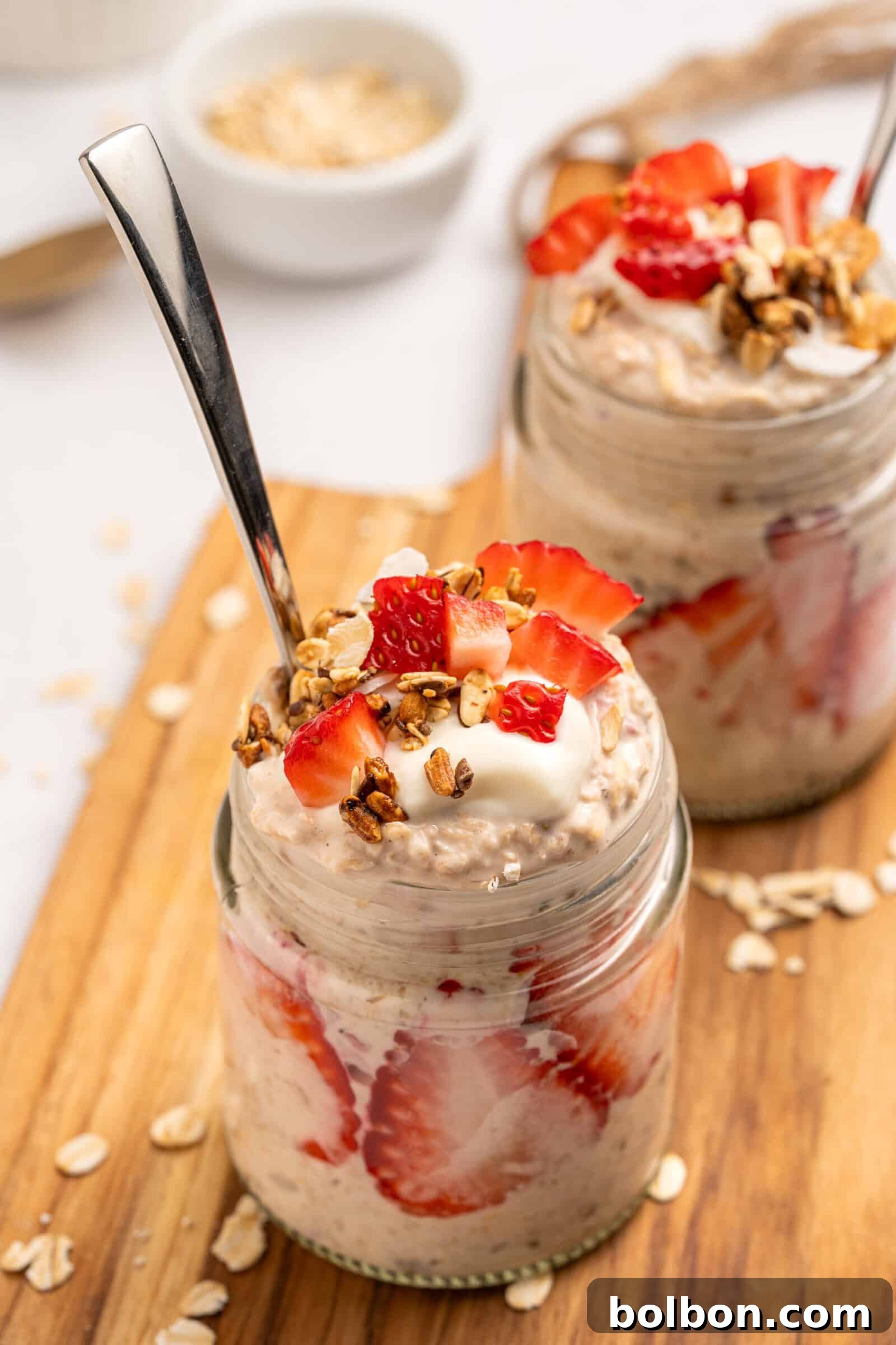Two glass jars filled, layered with sliced strawberries, topped with yogurt, granola, and fresh strawberries. Each jar has a spoon and sits on a wooden board with oats scattered around.