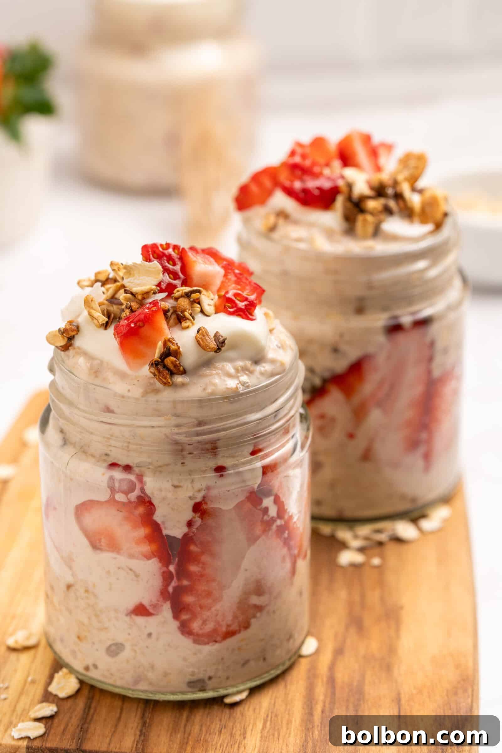 Two glass jars filled with overnight oats, layered with sliced strawberries and topped with creamy yogurt, fresh strawberry pieces, and granola, placed on a wooden board.