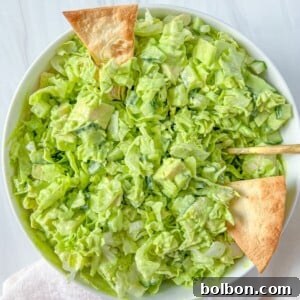 A bowl of chopped green salad with lettuce and cucumber, dressed lightly and topped with three triangular pita chips, sits on a white surface with a white napkin nearby.