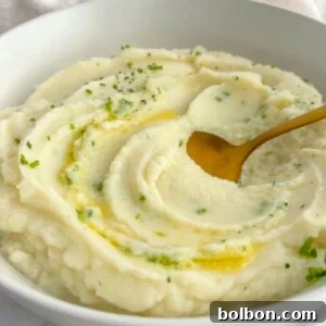A bowl of creamy mashed potatoes garnished with chopped herbs and a drizzle of melted butter, served alongside whole roasted cauliflower, with a gold spoon resting in the mash.