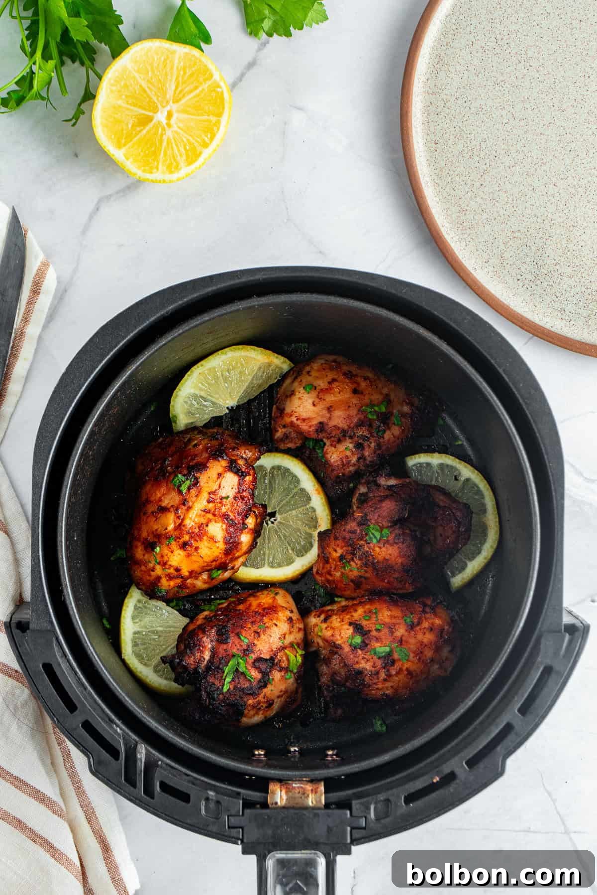 Perfectly Crispy Air Fryer Chicken Thighs 10 A top view of an air fryer basket containing four cooked chicken thighs garnished with lemon slices and herbs. A halved lemon, parsley, and a plate are placed nearby on the marble surface.