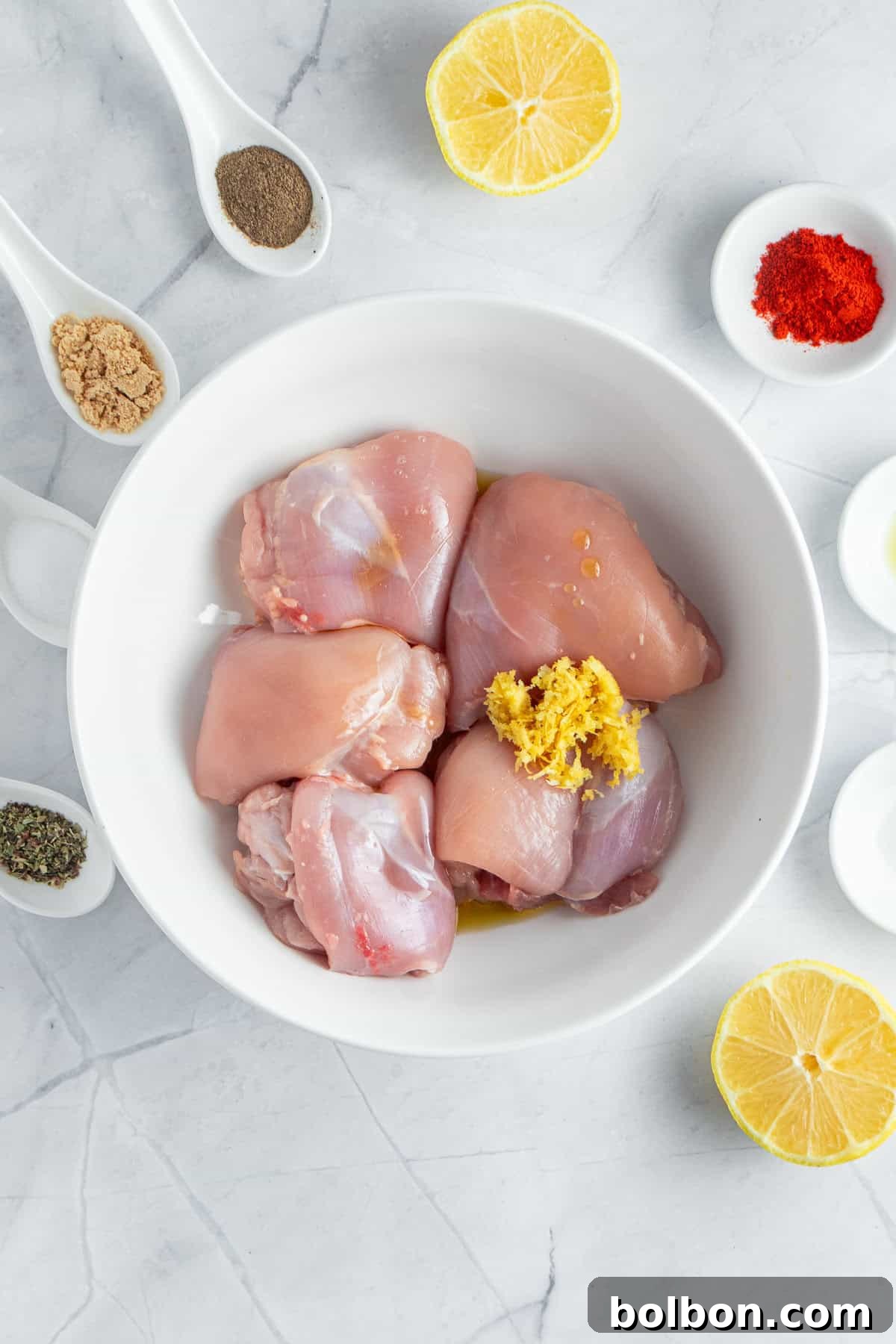 Perfectly Crispy Air Fryer Chicken Thighs 4 Chicken thighs in a white bowl with grated ginger on top, surrounded by lemon halves and small bowls containing ground spices, black pepper, salt, and red paprika, on a marble surface.