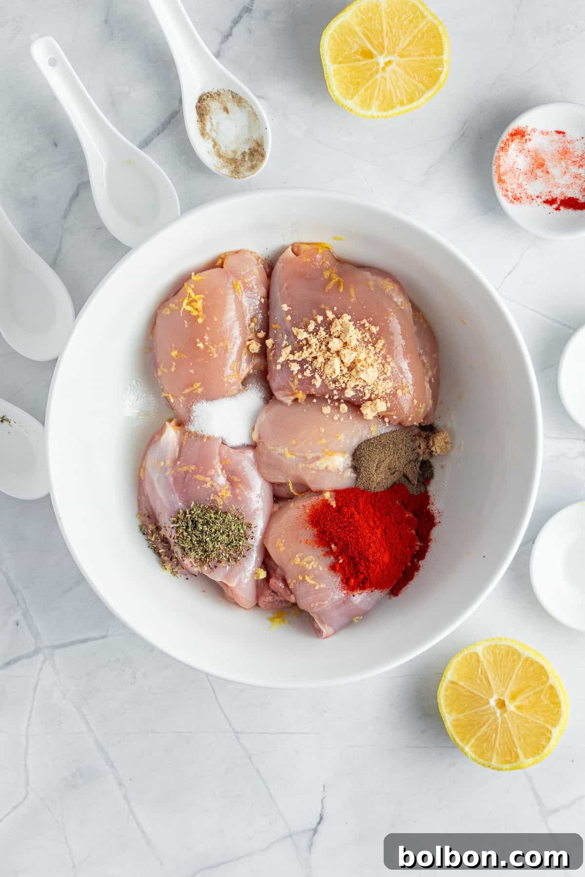 Perfectly Crispy Air Fryer Chicken Thighs 5 A white bowl filled with raw chicken pieces seasoned with spices, lemon zest, and herbs is surrounded by halved lemons and white spoons containing additional spices on a marble countertop.