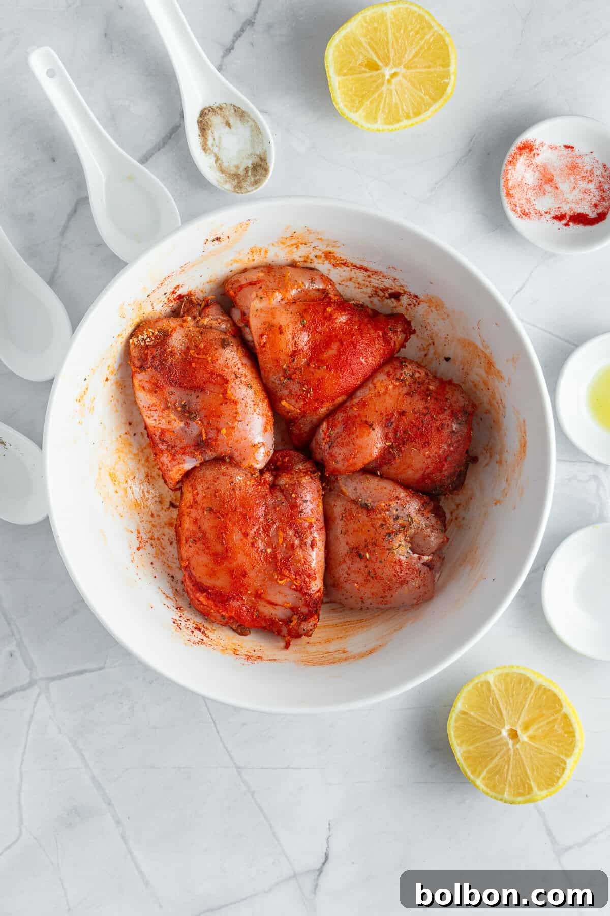 Perfectly Crispy Air Fryer Chicken Thighs 6 A white bowl contains raw chicken pieces marinated with spices. The bowl is surrounded by halved lemons and small white bowls filled with various spices on a marble surface. White measuring spoons also lie nearby.