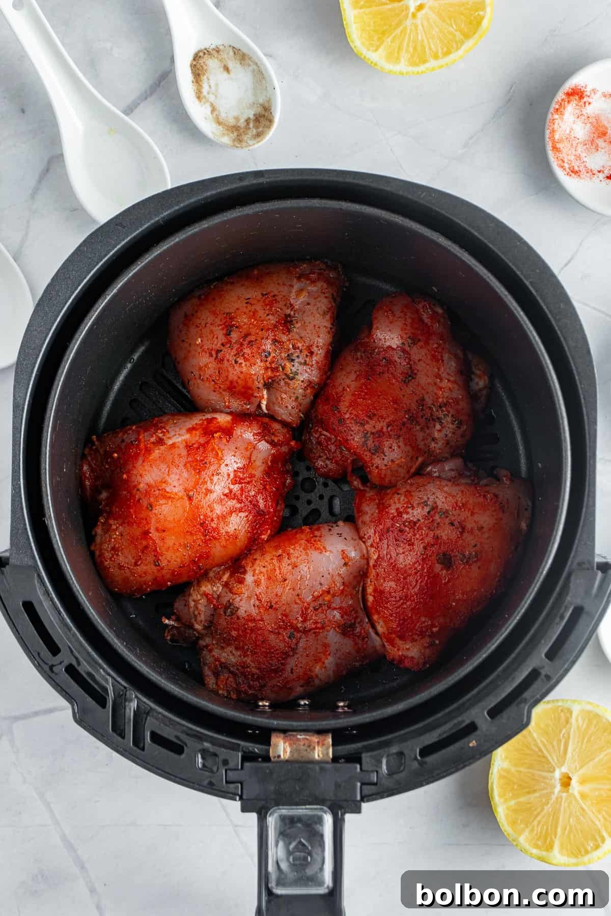 Perfectly Crispy Air Fryer Chicken Thighs 7 Air fryer containing seasoned and cooked chicken thighs. Slices of lemon and white spoons with spices are nearby on a marble surface.