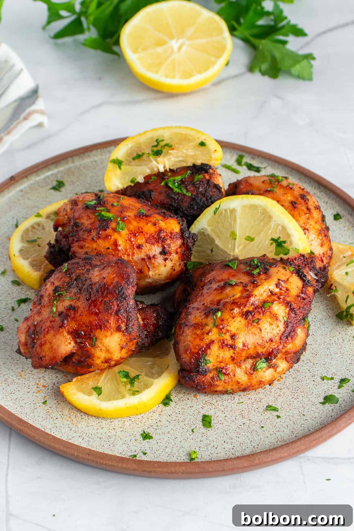 Perfectly Crispy Air Fryer Chicken Thighs 8 A plate of grilled chicken thighs garnished with lemon slices and fresh parsley. The chicken is browned and slightly charred, suggesting a flavorful, savory seasoning. In the background, there are cut lemons and parsley leaves.