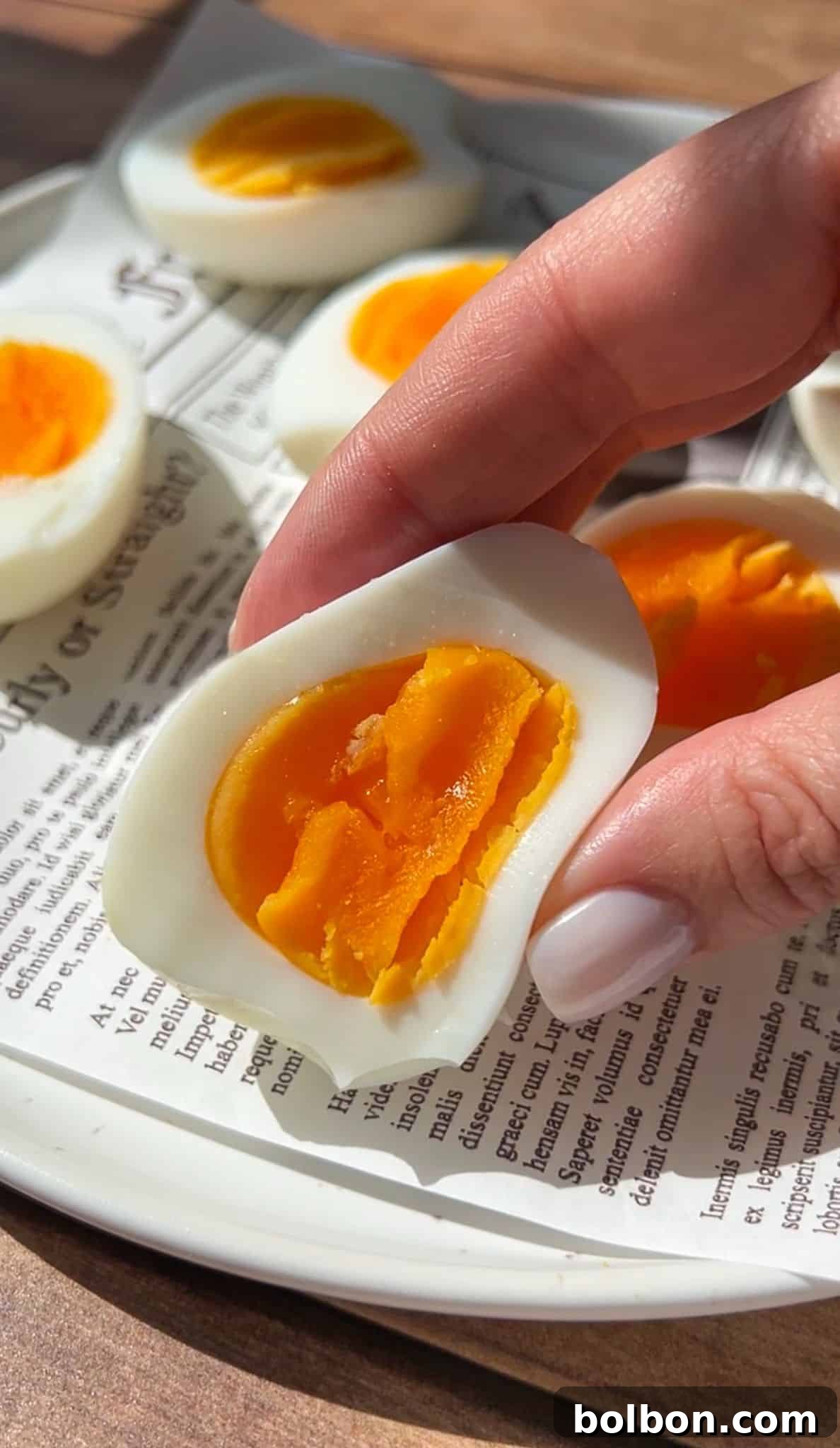 A hand holding half of a soft-boiled egg with a bright orange yolk; other halved eggs are on a plate lined with newspaper print.
