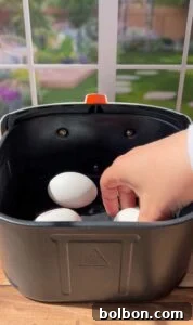 A hand places white eggs into the basket of a black air fryer, with a garden visible through a window in the background.