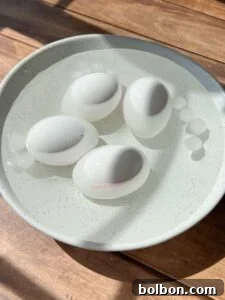 Four boiled eggs sit in a white bowl filled with water and ice cubes, placed on a wooden surface under bright sunlight.