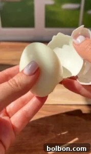 A person with light-painted nails is peeling the shell off a hard-boiled egg over a wooden surface, with sunlight shining and a blurred window in the background.